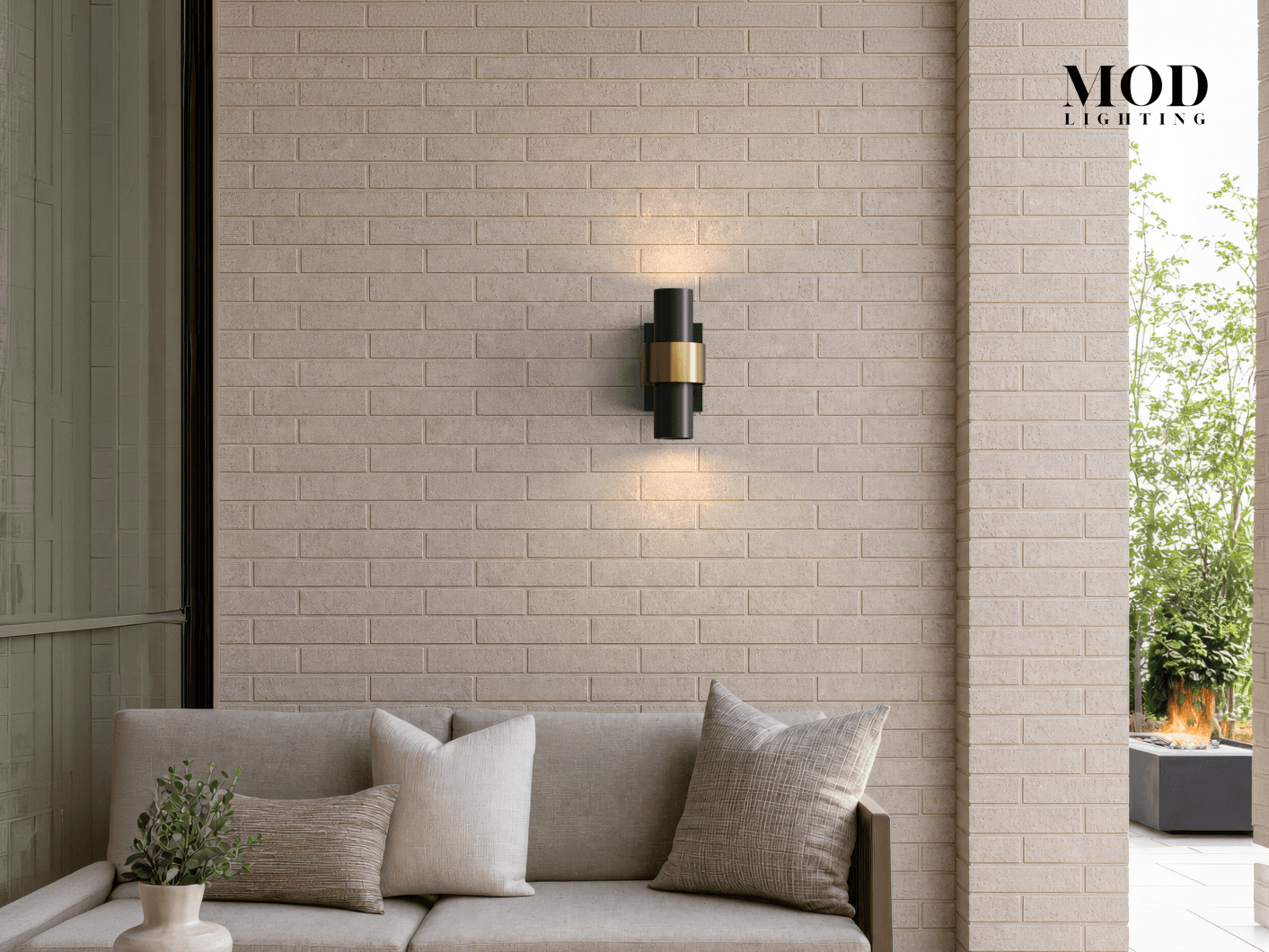 Stella black and brass outdoor sconce, banded cylinder glowing up and down on brick above a patio sofa